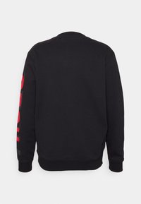 Sweatshirt noir avec un col rond, des manches longues et un texte rouge audacieux le long de la manche gauche. Le tissu semble doux et a une coupe décontractée.