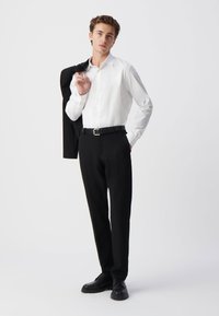 Witte lange mouw shirt, zwarte broek en zwarte schoenen. Zwarte jas over de linkerschouder gedrapeerd. Gladde stof met een aansluitend ontwerp.