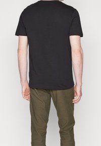 En man som bär en enkel svart t-shirt med kort ärm och olivgröna byxor, står vänd bort från kameran mot en ljus, enfärgad bakgrund.