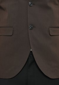 Brūns pielāgots uzvalka žakets ar melnu pogu, divām sānu kabatām un gludu faktūru, apvienots ar melnām biksēm apakšā.