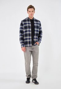 Blå och gräddfärgad rutig flanellskjorta med knäppning, bärs över en svart t-shirt, kombinerad med grå jeans och svarta sneakers.