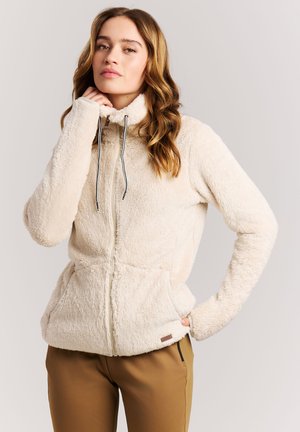 Frau trägt eine cremefarbene Fleecejacke mit hohem Kragen und Kordelzügen, kombiniert mit brauner Hose, posiert mit einer Hand am Hals.