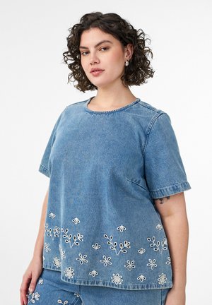 Kobieta o kręconych włosach nosi denimowy top i spodnie z białym kwiatowym haftem wzdłuż brzegu, stoi na białym tle.