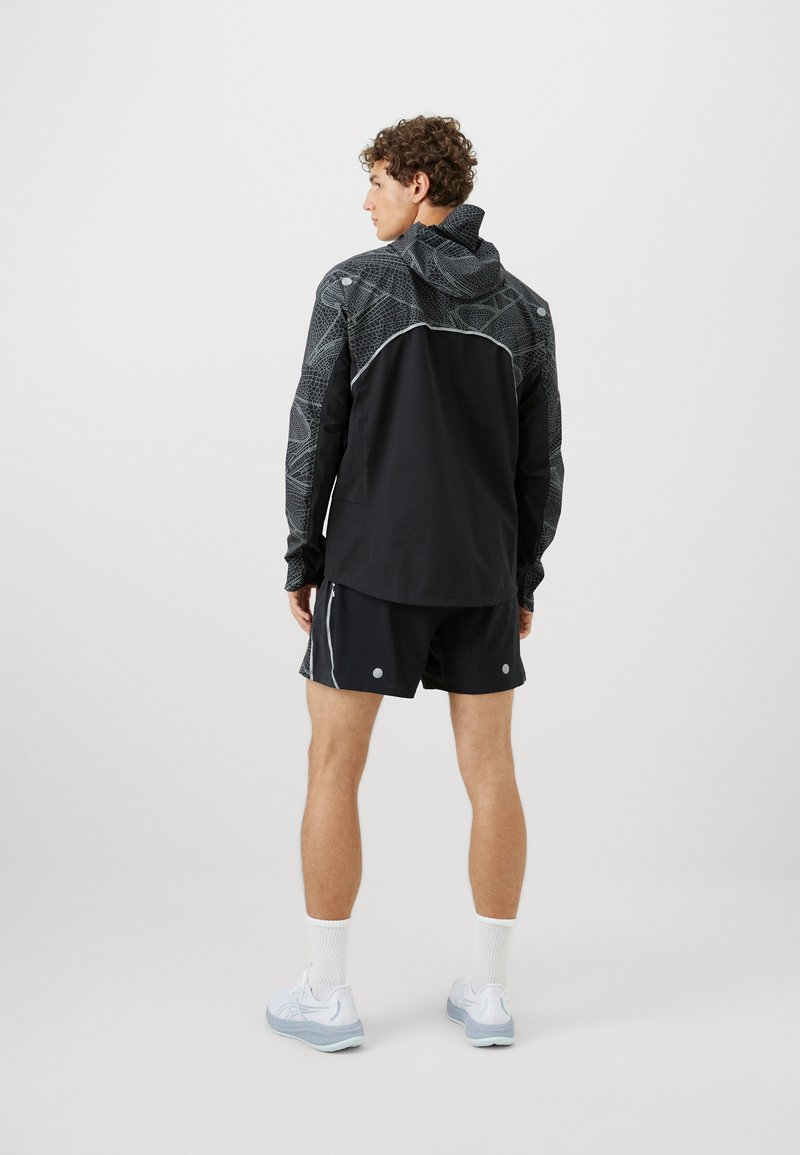 Chaqueta deportiva negra con hombros estampados, acentos blancos en contraste, combinada con pantalones cortos negros y zapatillas de correr azul claro.