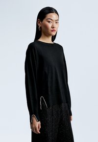 Černý pletený svetr s uvolněným střihem, žebrovaným lemem a dekoračními stříbrnými akcenty na manžetách. Nosený přes texturované černé kalhoty.