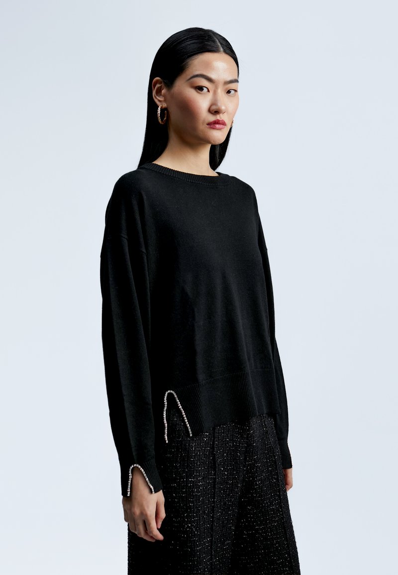 Černý pletený svetr s uvolněným střihem, žebrovaným lemem a dekoračními stříbrnými akcenty na manžetách. Nosený přes texturované černé kalhoty.