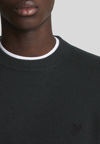 Suéter negro de cuello redondo, con un collar interior blanco y un pequeño logotipo bordado en el lado izquierdo. Tela suave y texturizada.