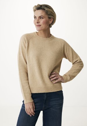Beige gebreide trui met een ronde halslijn en geribbelde manchetten, gestyled met donkerblauwe jeans. De textuur lijkt zacht en comfortabel.