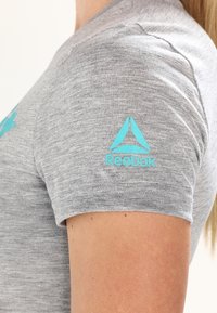 Camiseta de manga corta gris, hecha de una tela suave, con un logotipo de Reebok turquesa en la manga y acentos de diseño triangular.