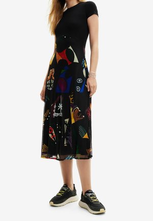 Kobieta ubrana w czarną bluzkę z krótkim rękawem i czarną spódnicę midi z kolorowymi abstrakcyjnymi wzorami, w połączeniu z czarno-białymi trampkami.