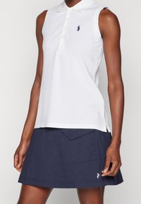 Bílý bezrukávový polo tričko s límečkem a třemi knoflíky, na kterém se nachází malé námořnické logo. V kombinaci s tmavě námořnickou sukní s přední kapsou.