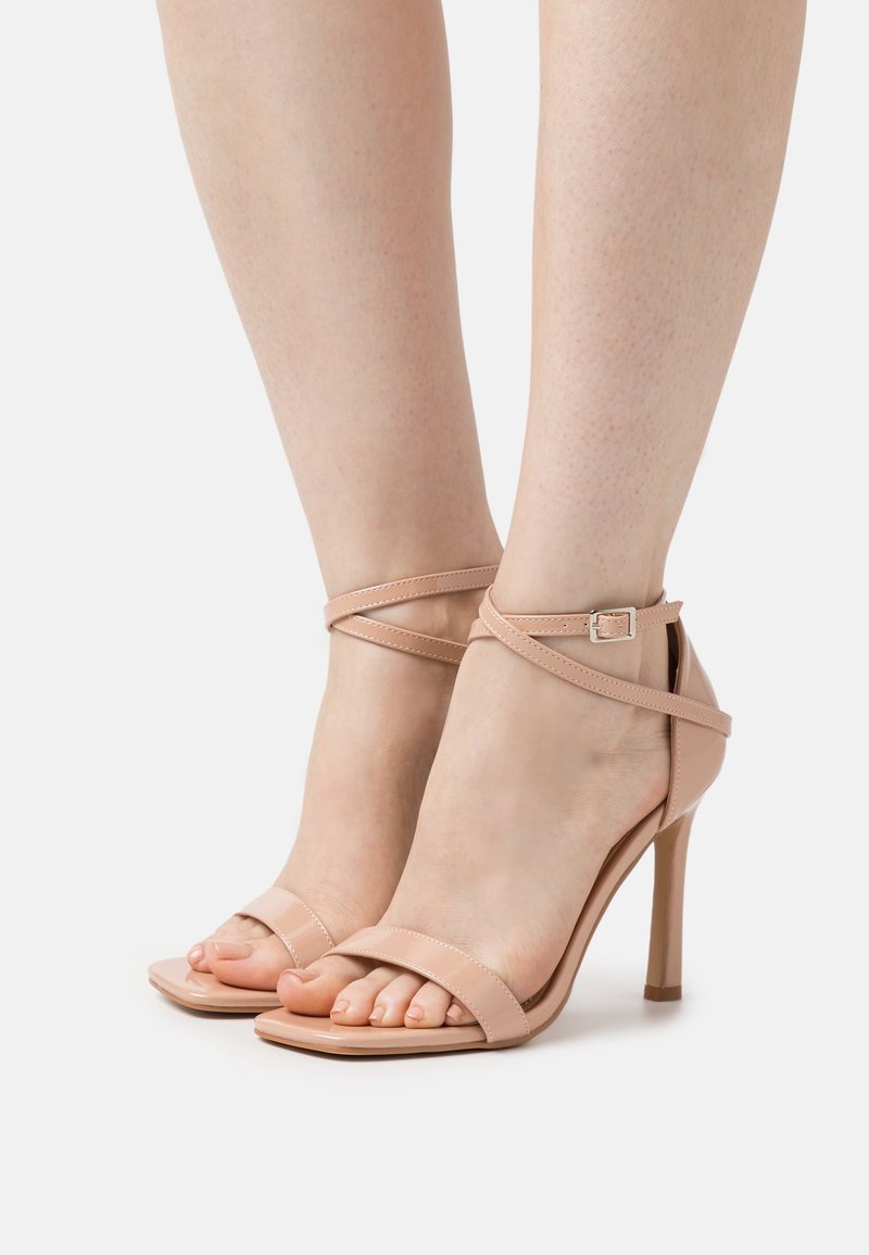 Pieds portant des sandales à talons hauts couleur nude avec des fines lanières cheville et orteils ouverts sur un fond blanc uni.
