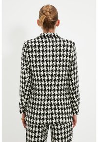 Blazer con motivo houndstooth in bianco e nero, caratterizzato da un design aderente, maniche lunghe e una chiusura con un solo bottone sul davanti.