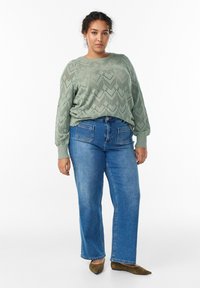 Gaiši zaļš adīts džemperis ar zigzag rakstu, apvienots ar augstām jostasvietām ziliem džinsiem un olīvu zaļās mokasīnām.