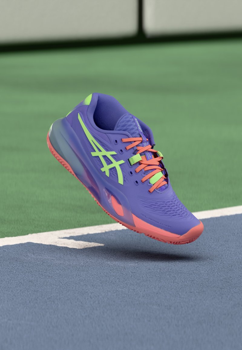 Μωβ αθλητικό παπούτσι με πράσινες και ρο ζ λεπτομέρειες, πλεκτό επάνω μέρος, υφή επιφάνειας και διαφανές πέλμα, τοποθετημένο πάνω από γαλάζιο γήπεδο.