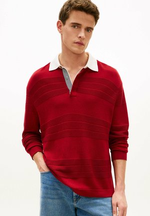 Jonge man die een rood rugbyshirt met lange mouwen met een witte kraag en een blauwe spijkerbroek draagt, staand met één hand in de zak tegen een witte achtergrond.