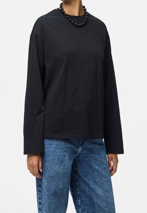 Chemise noire en coton à manches longues avec un col rond, associée à un jean denim bleu clair. Caractérisée par une texture lisse et une coupe décontractée. Collier de perles noires.