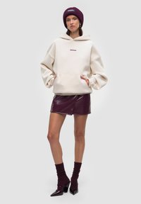 Hoodie oversize beige con tasca frontale e logo, abbinato a una mini gonna lucida color bordeaux, calze a costine bordeaux e tacchi neri.