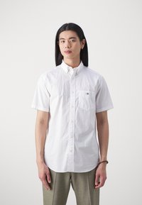 Man with long black hair wearing a white short-sleeve button-up shirt and grey pants, standing against a plain white background.