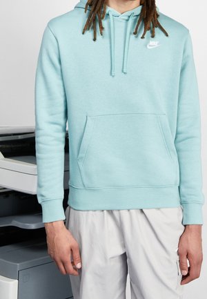 Mann mit langen Dreadlocks, der eine hellblaue Nike-Kapuzensweatshirt und hellgraue Hose trägt, steht neben einem Fotokopierer.