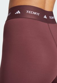 Leggings de sport bordeaux avec une texture lisse, dotés d'une large ceinture élastique avec "TECHFIT" et le logo adidas en blanc.