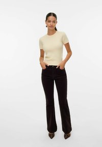 Top corto a righe beige chiaro con maniche corte abbinato a jeans a zampa di elefante marroni scuri, dotati di tasche frontali e scarpe punta marroni con dettagli metallici.