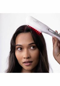 Femme utilisant une brosse capillaire LED StylPro blanche sur ses cheveux foncés près du cuir chevelu, sur un fond blanc uni.