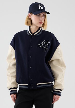 Mladá osoba nosí námornícku modrú a krémovú varsity bundu s nášivkou "NE" a námornícku čiapku New York Yankees, pozerá sa doprava na jednoliatom pozadí.