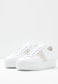 Pár bílých platformových tenisek s tkaničkami a béžovým pruhovaným logem NBKV po stranách, zobrazený na jednobarevném bílém pozadí.