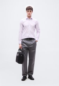 Νεαρός άνδρας όρθιος με το ένα χέρι στην τσέπη, φοράει ανοιχτό μωβ πουκάμισο με κουμπιά, γκρι παντελόνι, μαύρη ζώνη και παπούτσια, κρατώντας μαύρη τσάντα στο άλλο χέρι.