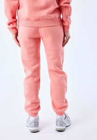 Koraalroze fleece joggers met geribbelde tailleband en boorden. Gecombineerd met witte en zilveren sneakers. Achteraanzicht, gladde textuur.