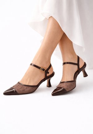 Benen met doorzichtige bruine slingback-hakken met spitse neus, bloemenborduurwerk en een gouden gesp, gecombineerd met een witte, vloeiende rok.