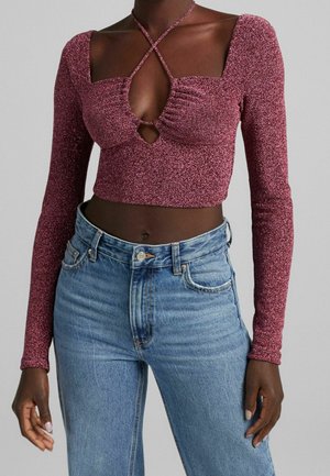 Personne à la peau foncée portant un crop top violet scintillant à manches longues avec un col croisé et un jean bleu clair taille haute.