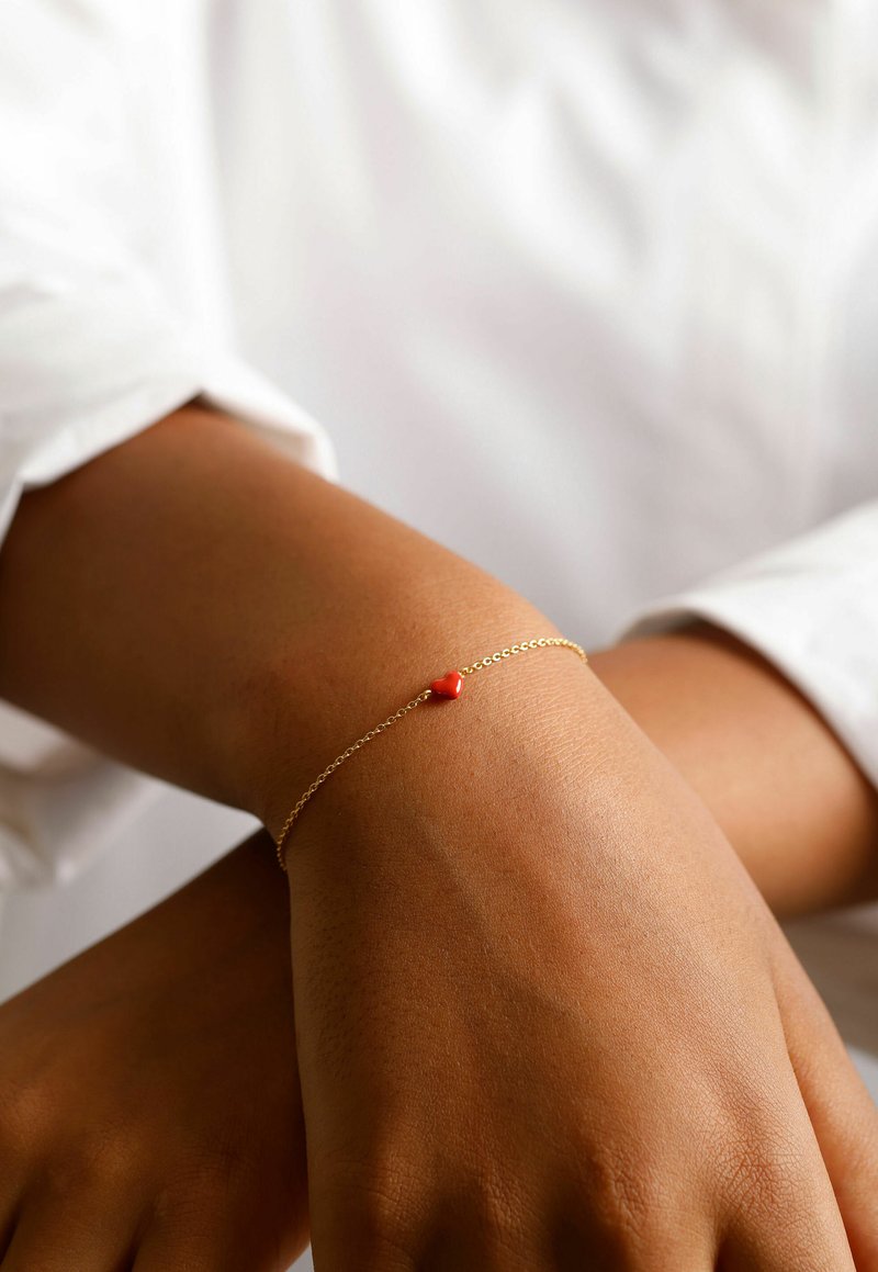 Bracelet en chaîne dorée avec un petit accent en forme de cœur rouge. La chaîne est délicate, et le cœur est lisse et arrondi, reposant sur le poignet.