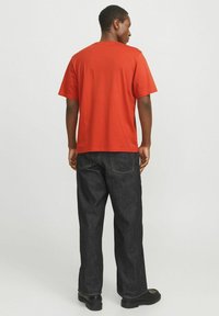 Rotes Baumwoll-T-Shirt mit lässiger Passform, kurzen Ärmeln und Rundhals-Ausschnitt, kombiniert mit dunklen Denim-Hosen und schwarzen Schuhen.