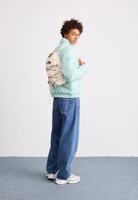 Valkoinen puffer-takki, rento istuvuus, käytetty löysien denimmien päällä. Valkoinen reppu mustilla yksityiskohdilla, joustava naru ja logo. Rentot kengät vihreillä yksityiskohdilla.
