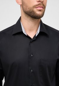 Zwarte overhemd met subtiele glans, voorzien van een contrasterende, patroonrijke kraag, knopen aan de voorkant en een borstzak.