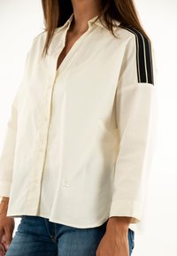 Camisa blanca de botonera con cuello, mangas de tres cuartos y acentos de rayas negras en los hombros. Logo bordado en el dobladillo.