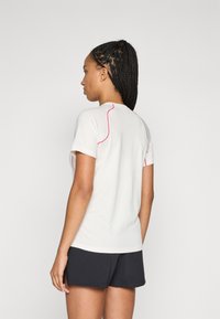 Kvindelig med fletning iført en hvid kortærmet skjorte med pink syninger og sorte shorts, stående med ansigtet mod venstre foran en ensfarvet baggrund.