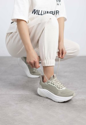 Zapatillas deportivas de color gris claro con parte superior de malla y suela blanca. Se están ajustando los cordones. El usuario lleva pantalones jogger de color claro.