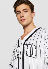Vit nätmesh baseballtröja med svarta vertikala ränder, med knäppning framtill och stora svarta bokstäver på bröstet.