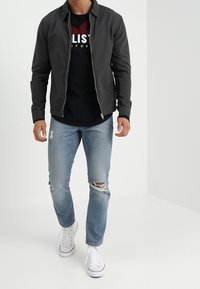 Men's black zip-up jacket over a black t-shirt, paired with light blue distressed jeans and white sneakers. Simple design and casual fit.