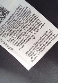 Bekleidungslabel, das den Stoffanteil mit 50 % Baumwolle und das Produktionsland Indien angibt, auf einem weißen Textiletikett in mehreren Sprachen gedruckt.
