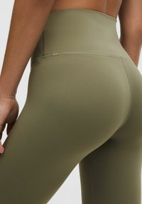 Közeli felvétel egy személyről, aki magas derekú olívazöld leggingset visel, a részletes varrás és szövettextúra látható a hát alsó részén és a csípőn.