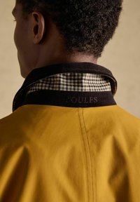 Man met kort krullend haar, gezien van achteren, die een mosterdgele jas draagt met een zwarte corduroy kraag en een geruite binnenkraag.