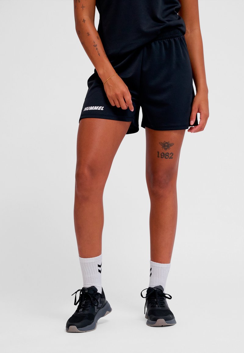 Shorts de sport noirs avec une taille élastique, arborant un logo blanc. Portés avec des baskets noires et des chaussettes blanches, mettant en valeur des tatouages sur les jambes.