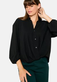 Black button-up blouse with a v-neck, billowing sleeves, and subtle texture. Combined with green pants, showcasing a relaxed fit.