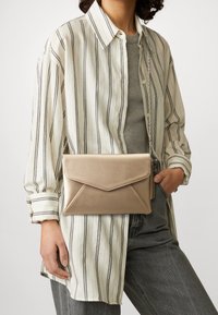 Metallisch goldene Clutch-Tasche mit einem Umschlag-Design, ausgestattet mit einem umklappbaren Deckel, einer Kettenriemen und einer glatten Textur. Getragen zu einem gestreiften Hemd.