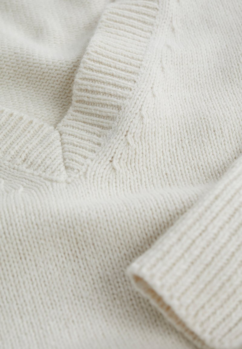Weißer Strickpullover mit V-Ausschnitt, geripptem Kragen und strukturiertem Körper. Nahaufnahme hebt den weichen Stoff und die detaillierte Naht hervor.