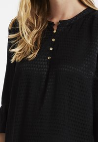 Svart blus med ett strukturerat mönster, rund hals och fem knappar i guldton framtill. Har långa ärmar med rynkade detaljer.